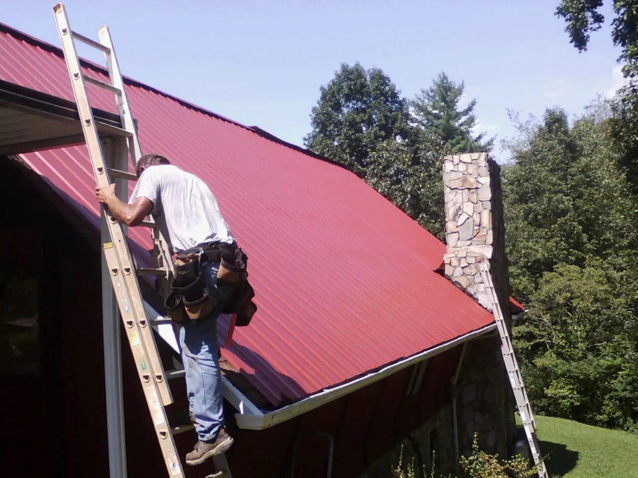 Roofer on a ladder beside a red metal roof and stone chimney — Soffit & Fascia Repair by Roofing Brothers in Minor Hill